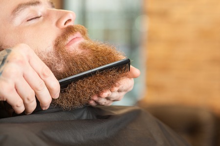 Close up of hands of professional barber combing beard of a man. The young bearded hipster is sitting with relaxation. He closed his eyes with pleasureの写真素材