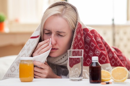 Sick blond girl has caught a cold. She is lying in bed and touching tissue to her nose. The lady closed her eyes with desperation. There are pills and vitamins on the tableの写真素材