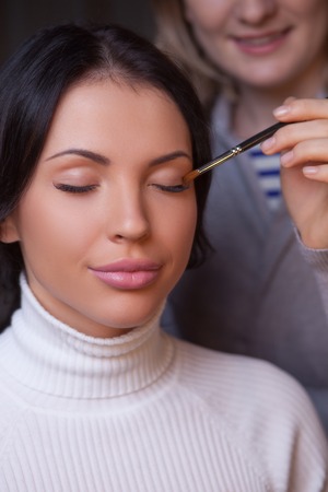 Attractive young woman is getting make-up. The female hand of artist is applying eyeshadow on her eyelid. She is standing and smiling. The model is sitting with closed eyes and smileの写真素材
