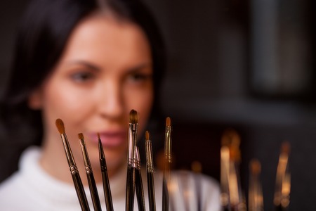 Attractive young woman is getting make-up. She is sitting and looking forward with joy. Focus on make-up brushの写真素材