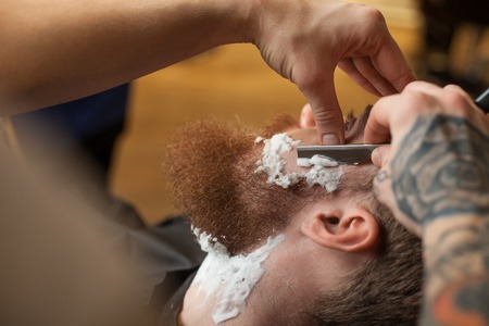 Close up of hands of barber shaving beard of the customer. He is touching a blade to human face. The man is sitting in chair with concentrationの写真素材