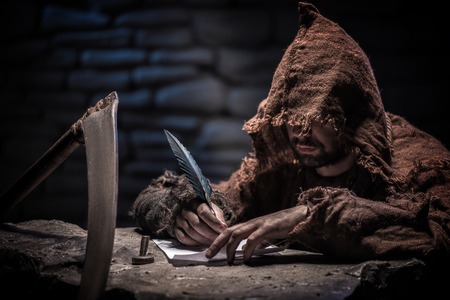 Grim Reaper is writing a list of his dead victims. The man is sitting near a scythe. He is holding a featherの写真素材