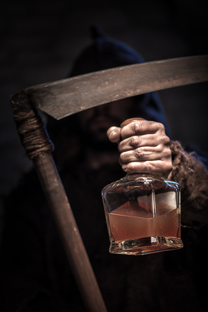 Dreadful death character is holding a scythe. He is stretching forward the bottle of alcohol drink with threatsの写真素材