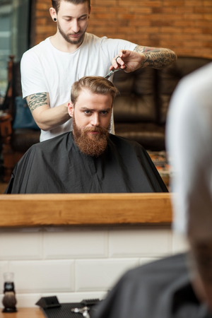 Professional young barber is cutting human hair with scissors and comb. He is standing and working with concentration. The bearded man is sitting and looking at mirror seriouslyの写真素材
