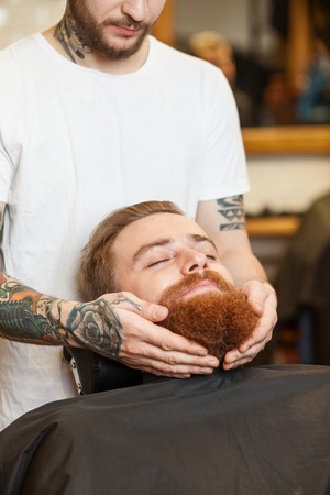 Close up of hands of skillful hairdresser touching the beard of man. He is preparing it for cutting. The young hipster is sitting and relaxing. His eyes are closedの写真素材
