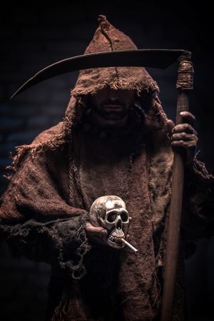 Grim Reaper is holding a scull smoking a cigarette. The man is standing in mantel. He is carrying a scythe with threatsの写真素材