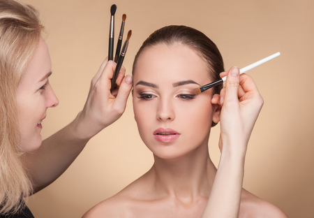 Skillful make-up artist is applying eye shadow on female eyelid. The woman is holding a brush and smiling. The young girl is sitting with relaxation. Isolated on brown backgroundの写真素材