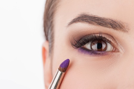 Close up of eye of cute young woman getting make-up at beauty salon. The brush is applying violet eyeshadow on her faceの写真素材