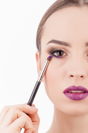 Close up of half of female face. The cheerful woman is looking forward with concentration. The make-up artist is applying violet eyeshadow under her eye with a brush. Isolatedの写真素材