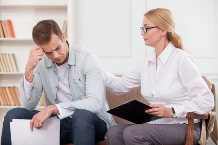 Attractive young man is visiting psychologist. He is crying with stress and holding papers. The woman is sitting near him on sofa. She is touching his back and trying to calm him downの写真素材
