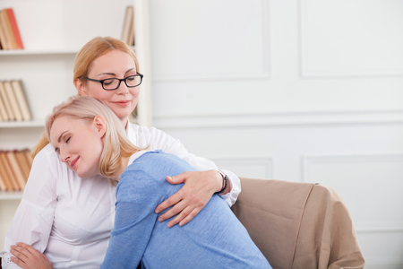 Experienced advisor is consulting young woman. They are sitting on sofa and embracing. The ladies are smilingの写真素材