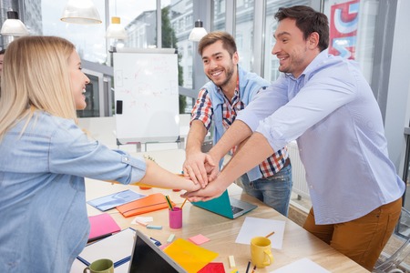 Cheerful young creative team is joining hands together. They are celebrating their unity. The men and woman are standing near the table in office.  They are smilingの写真素材