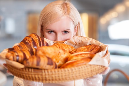 Cheerful young female baker is selling sweet pastry. She is standing and holding a tray of buns. The lady is looking at camera with desireの写真素材