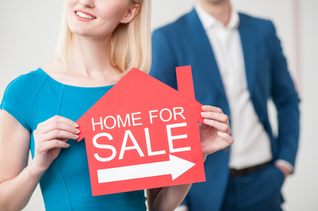 Home for sale. Close up of young two realtors advertising their business. The woman is showing a red placard and smiling. The man is standing on backgroundの写真素材