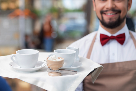 Close up of skillful young waiter standing and smiling. He is holding a tray with cups of coffeeの写真素材