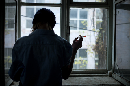 Frustrated young druggie is holding a syringe of heroine and preparing for injection. The woman is standing near the window. Focus on her backの写真素材