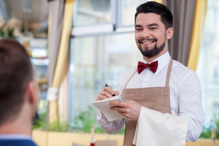 Skillful young waiter is serving a customer and smiling. He is standing and noting an order. The man is looking at the businessman with joyの写真素材