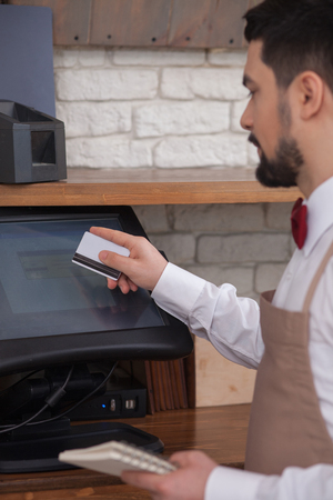 Cheerful young waiter is swiping a credit card through a machine. He is holding the notes and touching the screen with seriousnessの写真素材