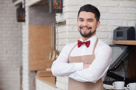 Attractive male cafe worker is standing near checkout machine. He is looking forward and smiling. The man is preparing an order. Copy space in left sideの写真素材