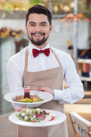 Cheerful male cafe is serving customers. He is standing and holding plate of salad. The man is looking forward and smilingの写真素材