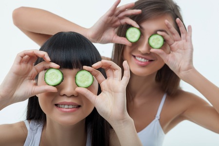 Attractive young women are covering their eyes with slices of cucumber. They are standing and smiling. Isolatedの写真素材