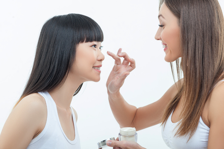 Pretty young women are standing and smiling. The Caucasian girl is applying cream on face of her asian friend. Isolated on backgroundの写真素材