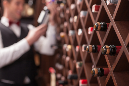 Cheerful young sommelier is choosing wine in cellar. He is smiling. Focus on bottles in shelfの写真素材
