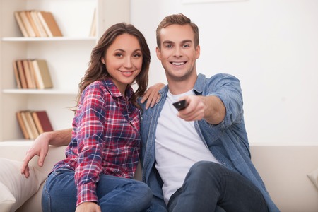 Beautiful young married couple is watching tv with joy. They are sitting on sofa and embracing. The...