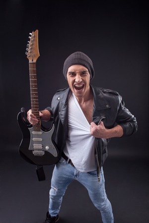 Portrait of wild guitarist expressing negative emotions. He is standing and trying to tear his clothing. The man is holding a guitar and shouting with anger.の写真素材