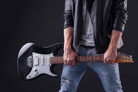 Close up of professional guitarist holding a guitar with strength. He is standing in jeans and leather jacket. Isolatedの写真素材