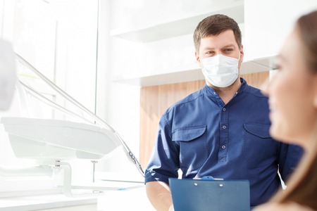 Handsome dentist is standing and holding a folder. He is analyzing the results of curing with concentration. The woman is sitting in chair and smiling. Copy space in left sideの写真素材