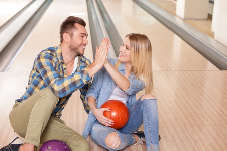 We are the great team. Portrait of beautiful young men and woman giving high five in bowling club. They are sitting on floor and smiling. The blond girl is holding a ballの写真素材