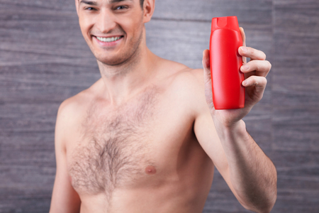 Portrait of handsome young man presenting new shampoo. He is standing in the bathroom with naked torso. The macho is holding a bottle and smilingの写真素材