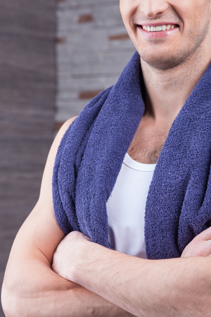I want to be clean and fresh. Cheerful young man is standing in a bathroom with a towel on his shoulder. He is smiling. The man crossed his arms confidentlyの写真素材