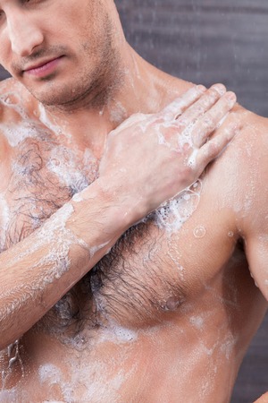Handsome young man is washing his body with soap. He is standing and touching his shoulder with relaxationの写真素材
