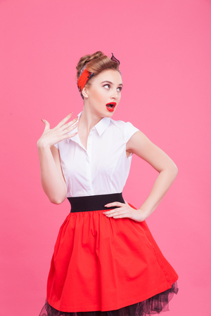 Portrait of beautiful young woman standing and posing in retro dress. She is looking aside with amazement and waving her arm near the face. Isolatedの写真素材