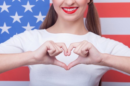 Beautiful girl is expressing her love to United States of America. She is standing and making heart with her arms. The lady is smiling happilyの写真素材