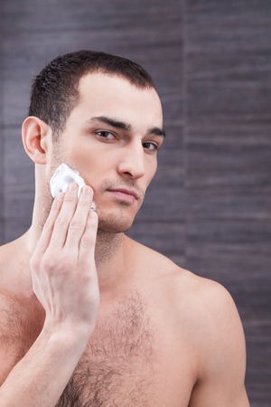 Portrait of handsome young man applying shaving foam on his chin. He is standing and looking at camera with confidenceの写真素材
