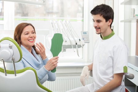 Portrait of mature woman sitting in medical chair after treatment. She is holding a mirror and looking at her white teeth with satisfaction. The dentist and lady are smilingの写真素材