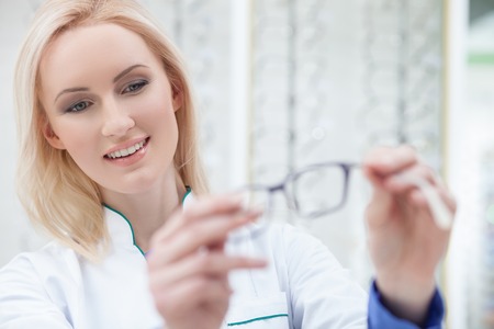 Waist up portrait of professional young optician serving the customer in shop. The woman is standing and holding eyeglasses. She is smilingの写真素材