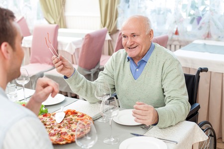 Old man has a dinner with his young son in restaurant. They are talking with joy. The men are smiling. The father is sitting on the wheelchairの写真素材