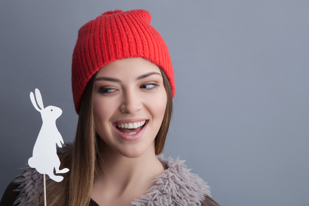 Portrait of attractive woman holding a little figure of a rabbit on the stick. She is playing with it and laughing. The lady is wearing a hat. Isolated and copy space in right sideの写真素材