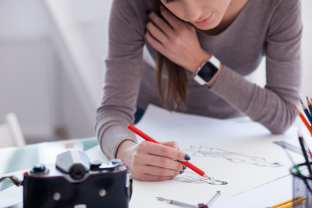 Pretty young professional photographer is drawing sketch on the blueprint. The woman is standing near the desk and smiling. There is a camera on the tableの写真素材