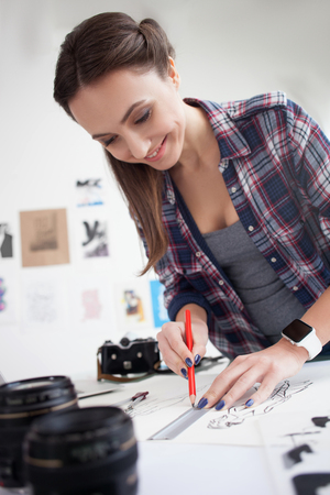 Skillful young photographer is drawing sketches with joy. The woman is standing and the table and...