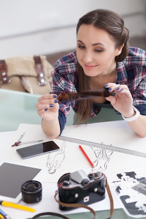 Attractive female photographer is taking a view of an old photographic film. She is sitting at the table near sketches and camera. The woman is smilingの写真素材
