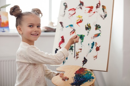 Portrait of cute small girl learning to paint at home. She is standing near the canvas and holding the palette. The kid is looking at camera and smilingの写真素材