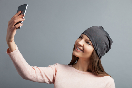 Pretty young woman is making selfie. She is standing and holding a mobile phone. The lady is posing and smiling. Isolatedの写真素材
