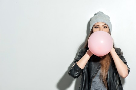 Beautiful young woman is inflating the pink balloon. She is standing and looking forward happily. The lady is wearing a hat and black leather jacket. Isolated and copy space in left sideの写真素材