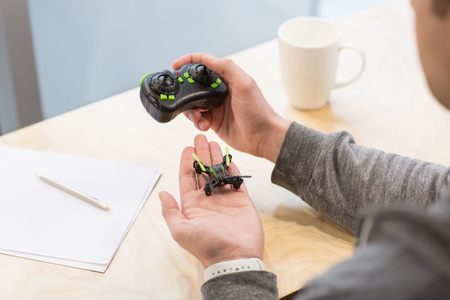Skillful young freelancer is designing a new quadrocopter. He is sitting at the desk near documents. The worker is holding a small drone and remote controllerの写真素材