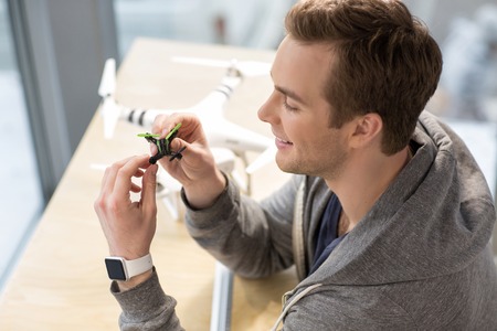 Cheerful young man is working with small quadrocopter. He is sitting and smiling. The freelancer has a smart watch on his armの写真素材